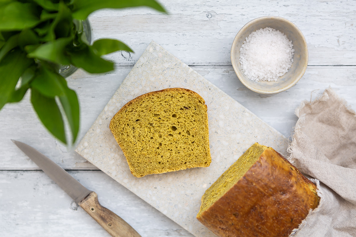 Angeschnittener Laib Bärlauch-Brioche auf einem Schneidebrett, daneben steht ein Glas mit frischem Bärlauch.